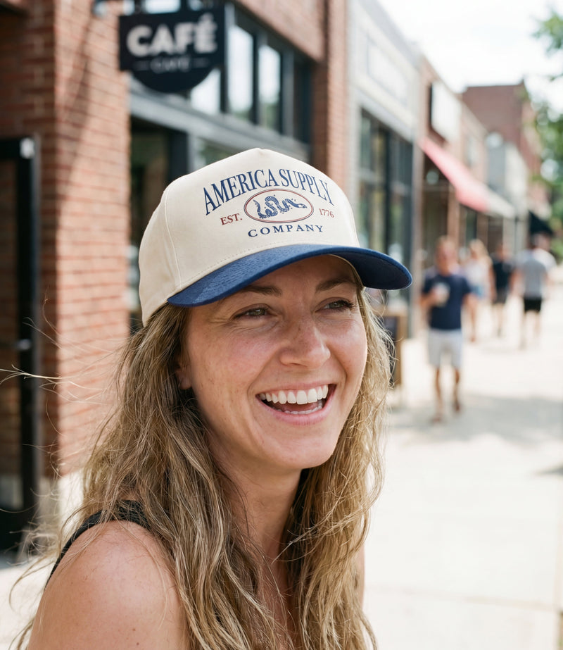 The Alex Jones Store - Smiling woman wearing tan and navy America Supply Co. 'Est. 1776' Trucker Hat with rattlesnake logo on city sidewalk
