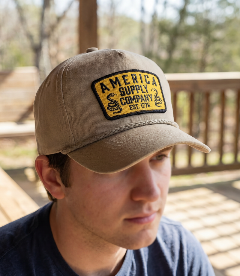 The Alex Jones Store - Young man wearing tan America Supply Company snake patch hat with navy shirt seated on a wooden porch outdoors