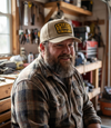 The Alex Jones Store - Bearded man smiling in tan America Supply Company patch cap and plaid flannel shirt inside a wood-paneled garage workshop
