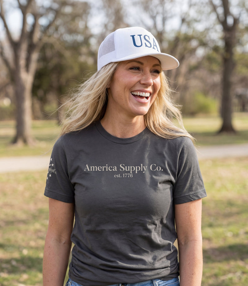 The Alex Jones Store - Blonde woman laughing outdoors in gray America Supply Co. 'Est. 1776' tee and white USA trucker hat