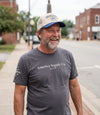 The Alex Jones Store - Bearded man smiling on sidewalk wearing gray America Supply Co. 'Est. 1776' tee and America Supply Co. trucker hat