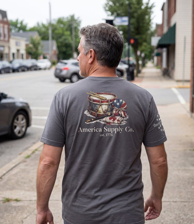 The Alex Jones Store - Man walking down street wearing gray America Supply Co. 'Est. 1776' tee, back view showing drum and flag print