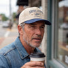 The Alex Jones Store - Man in cream and navy America Supply Co trucker hat with 'Est. 1776' rattlesnake logo, holding coffee on a city street
