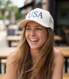 The Alex Jones Store - Smiling brunette woman wearing white 'USA' Premium Trucker Hat with navy serif USA print, outdoor cafe setting